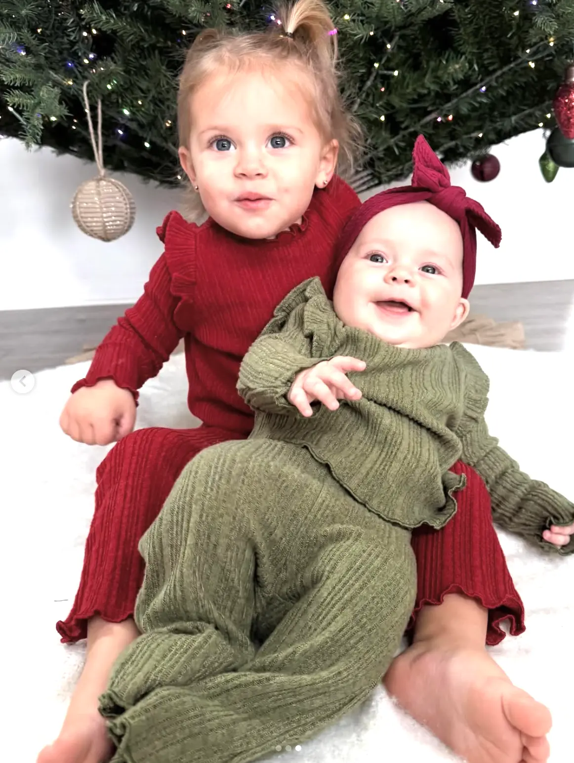 Two sisters in red and green knits sitting under a lit Christmas tree