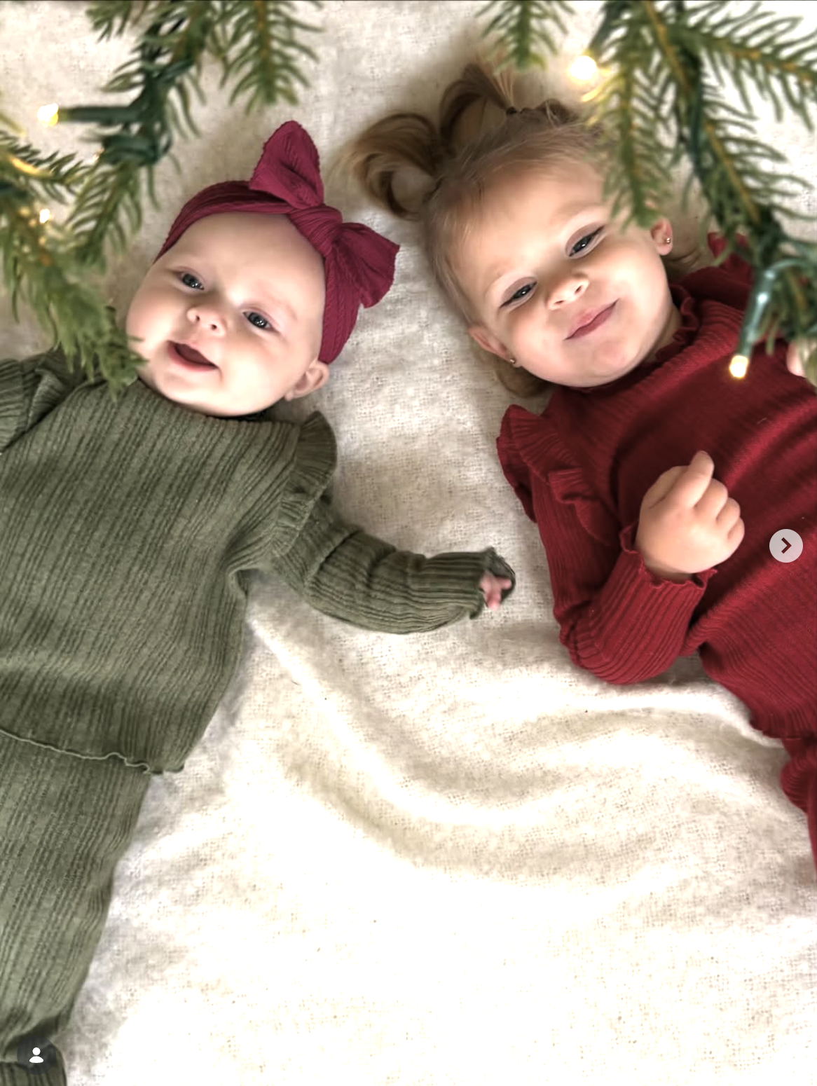 Two babies in red and green outfits lying among greenery and fairy lights